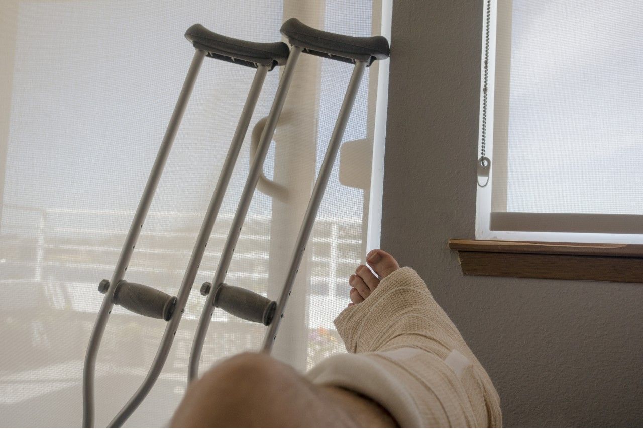 Disabled Injured Person With Sprained or Broken Ankle or Foot Sits Inside With Crutches Looking Outside the Sliding Glass Door Window on a Sunny Day.