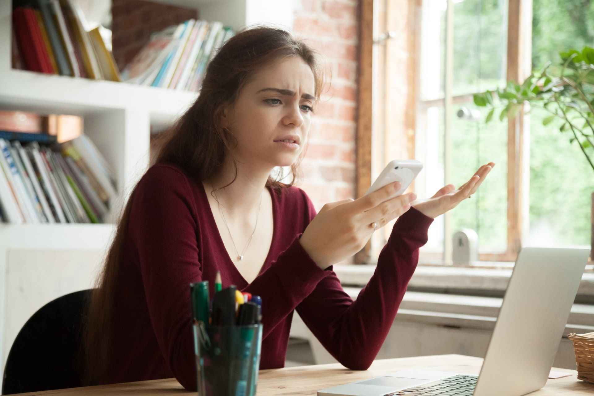 Annoyed woman in a maroon shirt looks at her smartphone while sitting at a desk with a laptop