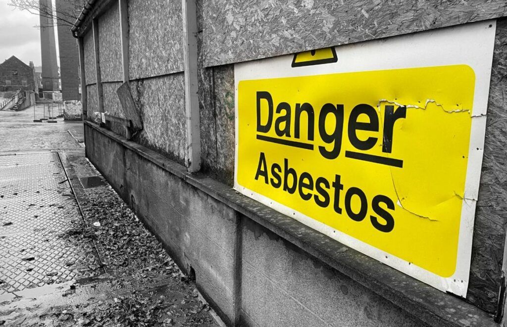 A white-and-yellow "Danger Asbestos" sign on a plywood wall outdoors, background in black and white 